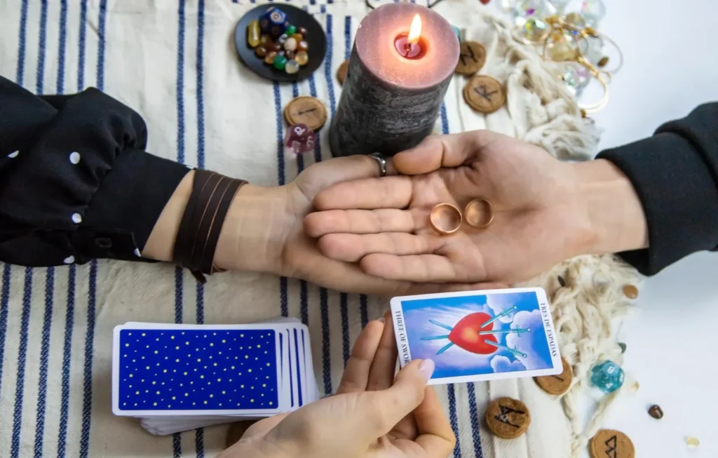 Una sesión de tarot enfocada en la reconciliación de pareja. En la imagen, dos personas sostienen sus manos abiertas donde descansan dos anillos de oro, simbolizando el compromiso y el deseo de unión. En primer plano, una mano sostiene la carta del tarot "Tres de Espadas" (corazón atravesado por espadas), representando el dolor de la separación que se busca superar. El escenario incluye una vela encendida, runas de madera y piedras energéticas sobre un mantel rústico, ideal para ilustrar rituales y consultas sobre cómo regresar con un ex.