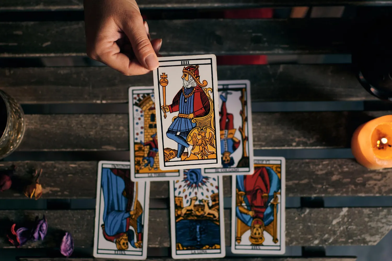 Una toma cenital de una lectura de tarot mística sobre una mesa de madera oscura y rústica. Una mano sostiene en el aire la carta del tarot "IIII. EL EMPERADOR" (del Tarot de Marsella), que es la central y está en posición vertical. Debajo de ella hay una tirada de cartas en cruz, todas invertidas, incluyendo (de izquierda a derecha y arriba a abajo) La Torre invertida, El Colgado invertido, El Diablo invertido y La Luna invertida. A los lados hay una vela encendida y pétalos secos esparcidos, creando un ambiente de adivinación.